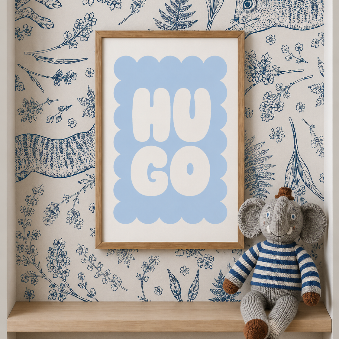 A framed nursery art print with the word 'HUGO' in white letters on a light blue background, styled on a wooden shelf against blue-and-beige illustrated wallpaper with a stuffed elephant toy.