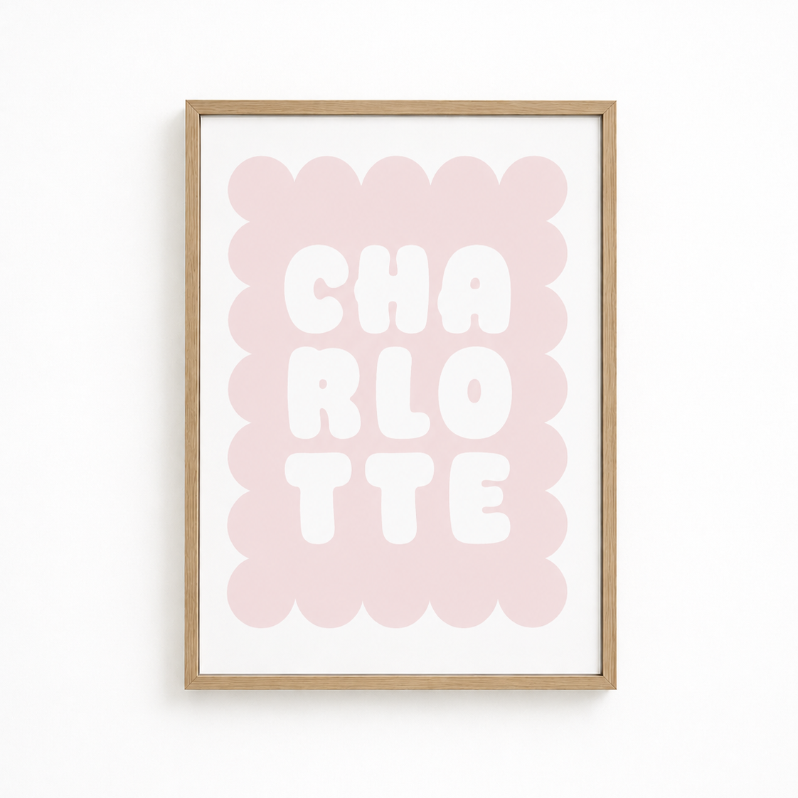 A framed typographic wall art print featuring the word 'CHARLOTTE' in large white letters over a soft pink scalloped rectangle, mounted in a light natural wood frame against a white wall.